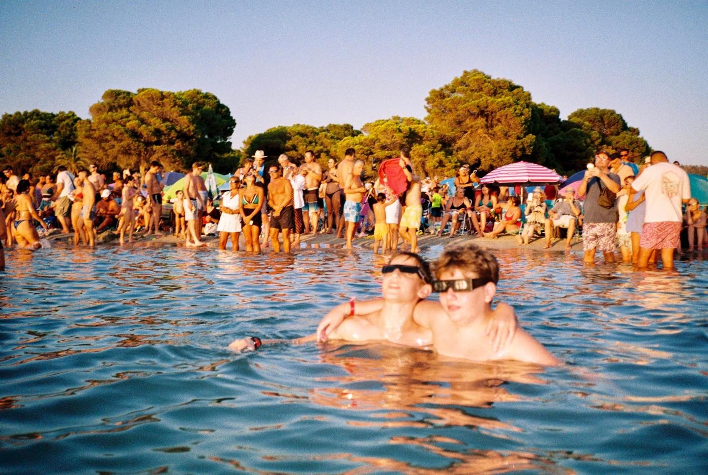 Two boys wearing eclipse glasses float in the water while a crowd of people gather on a beach in the background.