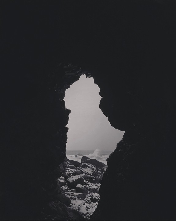 A view of the North Atlantic Ocean with waves crashing on rocks is seen through a dark cave opening.