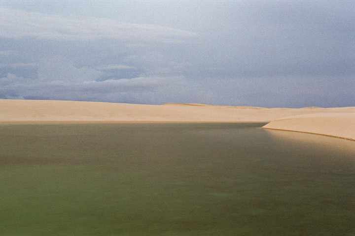A still body of water is surrounded by sand dunes under a cloudy sky.