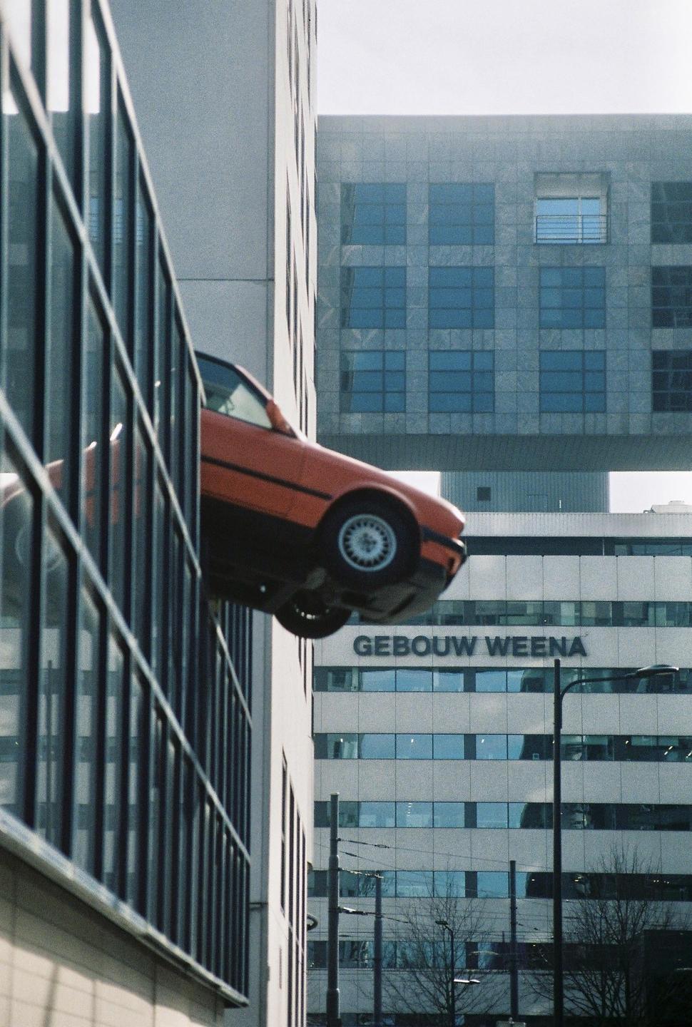 An orange car is suspended vertically on the side of a modern building with glass windows.