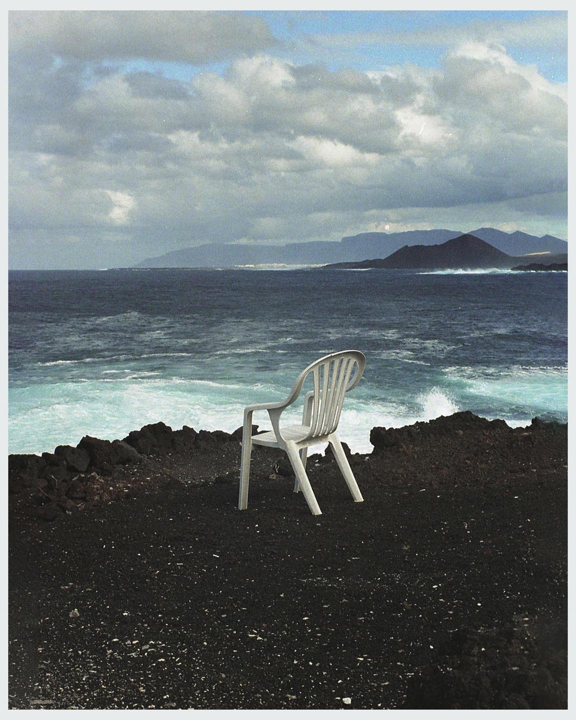 A white plastic chair sits on black volcanic rock overlooking a rough ocean with mountains in the distance under a cloudy sky.