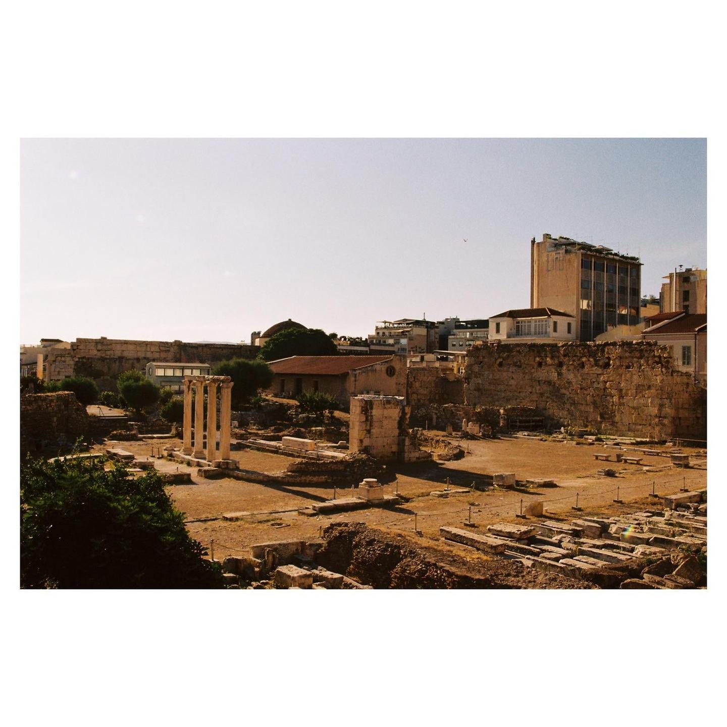 Ancient ruins with tall columns are visible in the foreground, with modern buildings and a clear sky in the background.