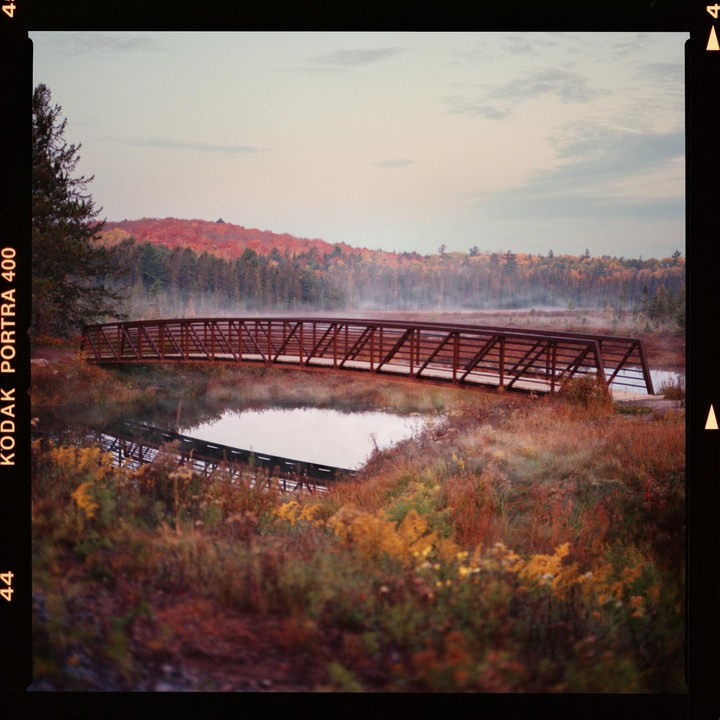 A metal bridge crosses a body of water with autumn foliage and fog in the background.