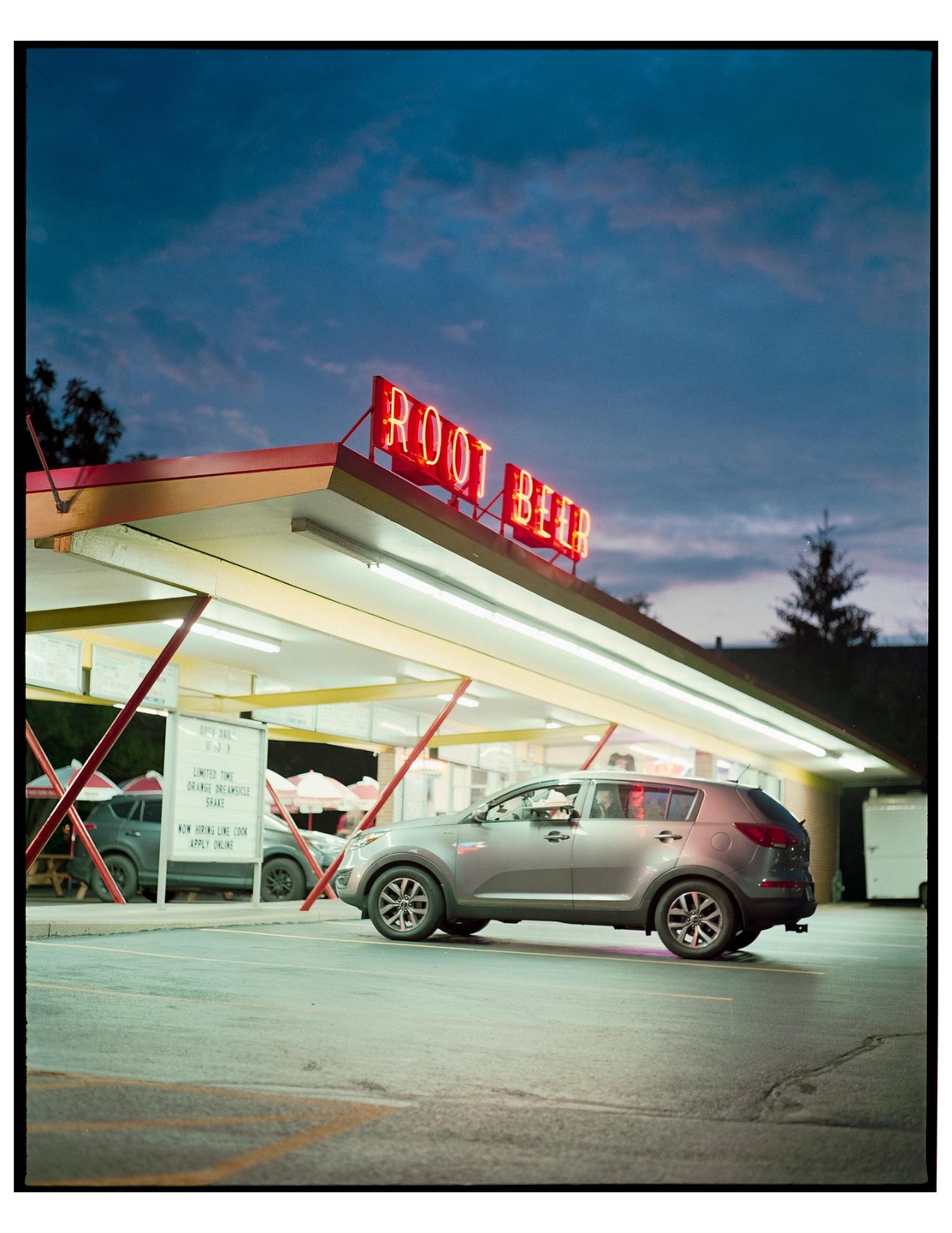 A gray vehicle is parked under a drive-in canopy with a neon "ROOT BEER" sign against a twilight sky.