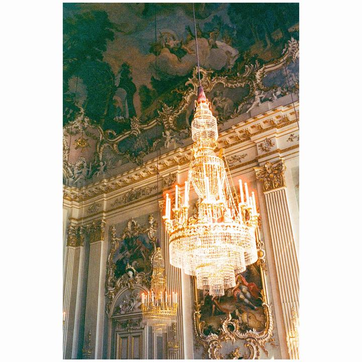 A large, ornate crystal chandelier hangs from a highly decorated ceiling in a grand room.