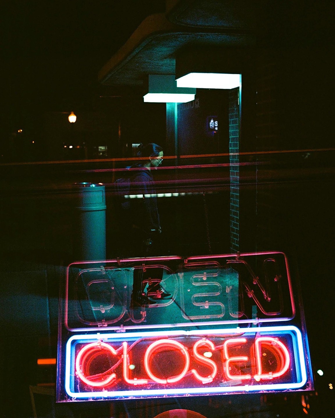 A neon sign for an open and closed business is visible at night with a person walking in the background.
