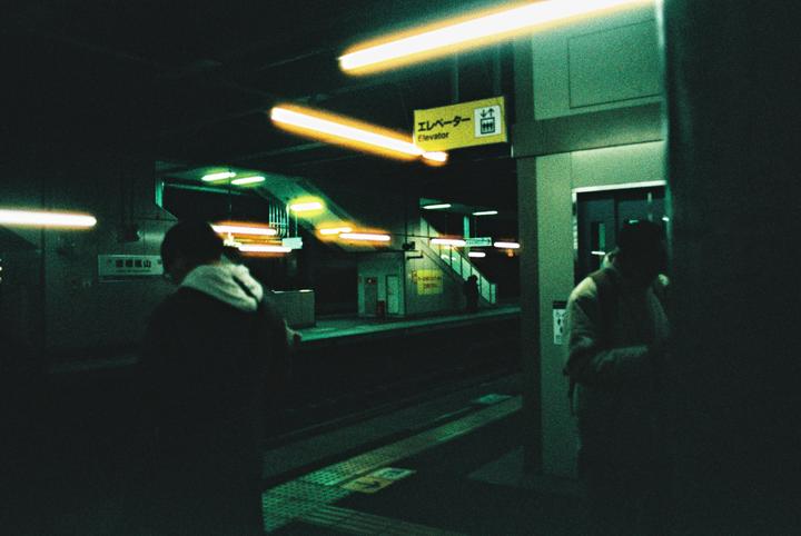 Two people stand on a dark subway platform with bright yellow and green lights.