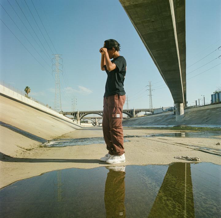 A boy stands in a concrete riverbed with a bridge overhead and power lines in the distance.