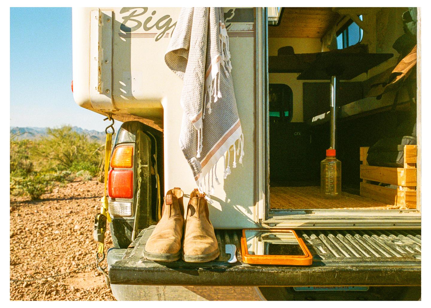 A pair of boots and a mirror sit on the tailgate of a truck with a camper shell in a desert landscape.