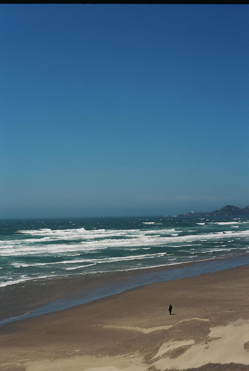 A lone figure stands on a wide sandy beach with waves crashing in the background and a distant lighthouse on a headland.