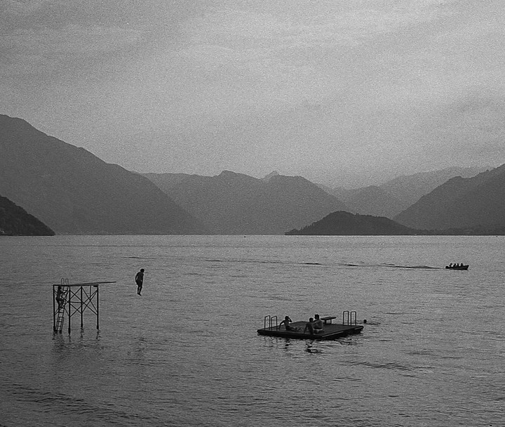 A person jumps from a diving platform into a lake surrounded by mountains, while a boat and a floating platform with people are also visible.