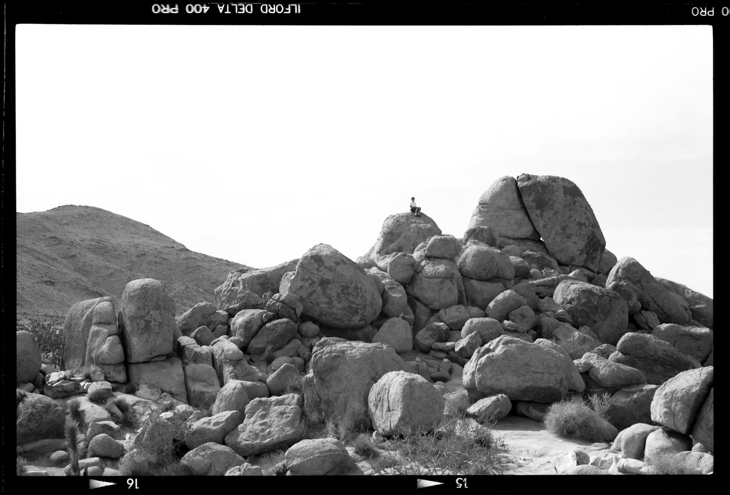 A man sits atop a large rock formation in a desert landscape with a hill in the background.