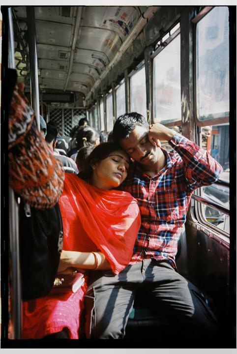 A couple is sleeping on a bus with other passengers in the background.