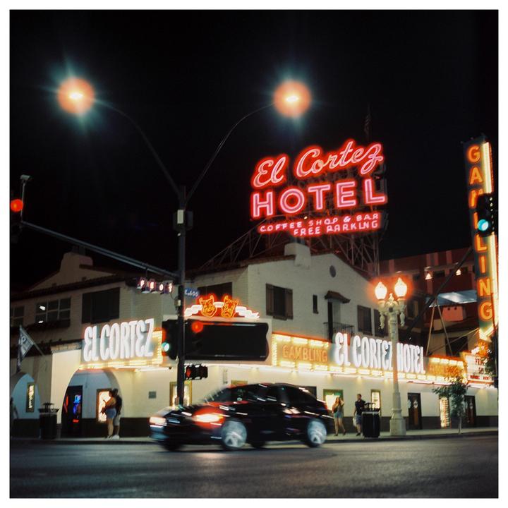 A black car with motion blur drives past the El Cortez Hotel at night, illuminated by neon signs and streetlights.