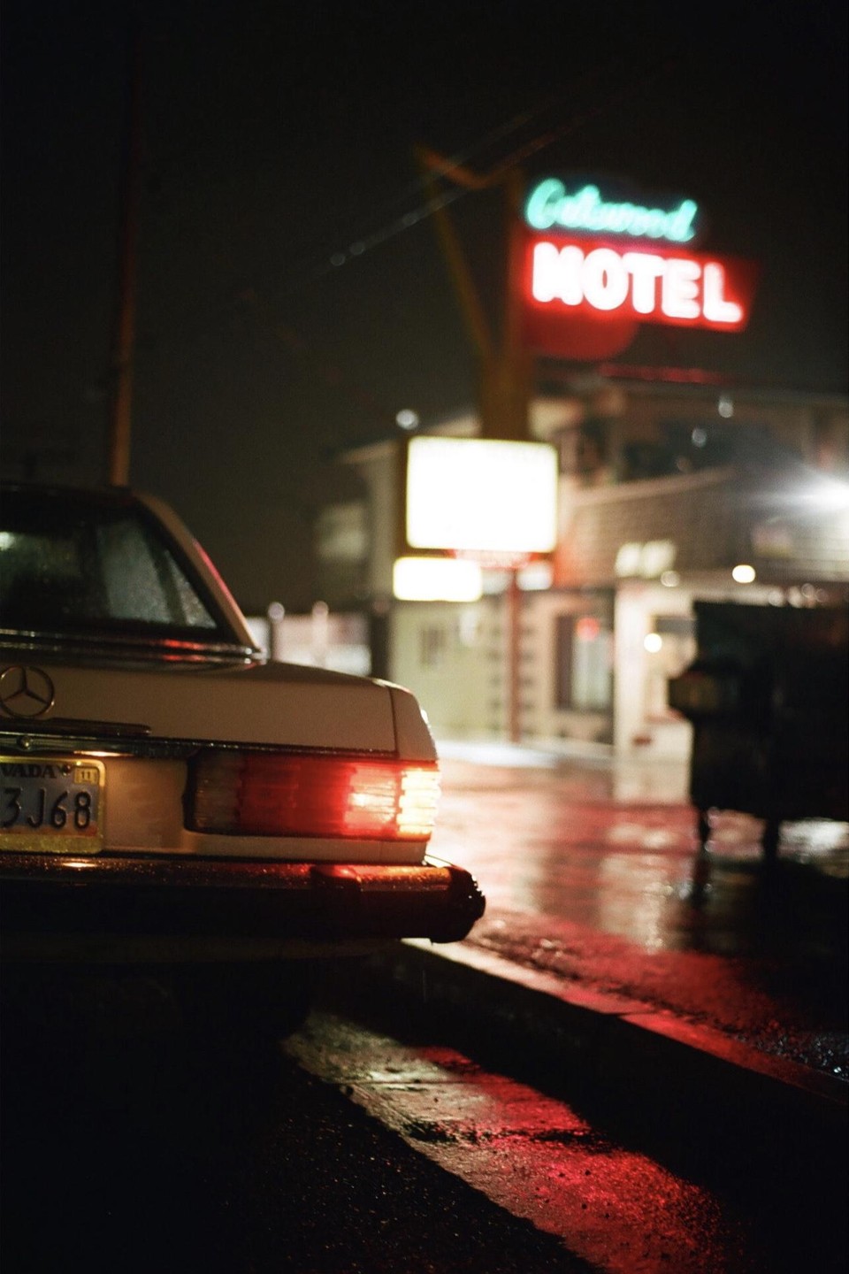 The rear of a white car is parked on a wet street at night, with a neon motel sign visible in the background.