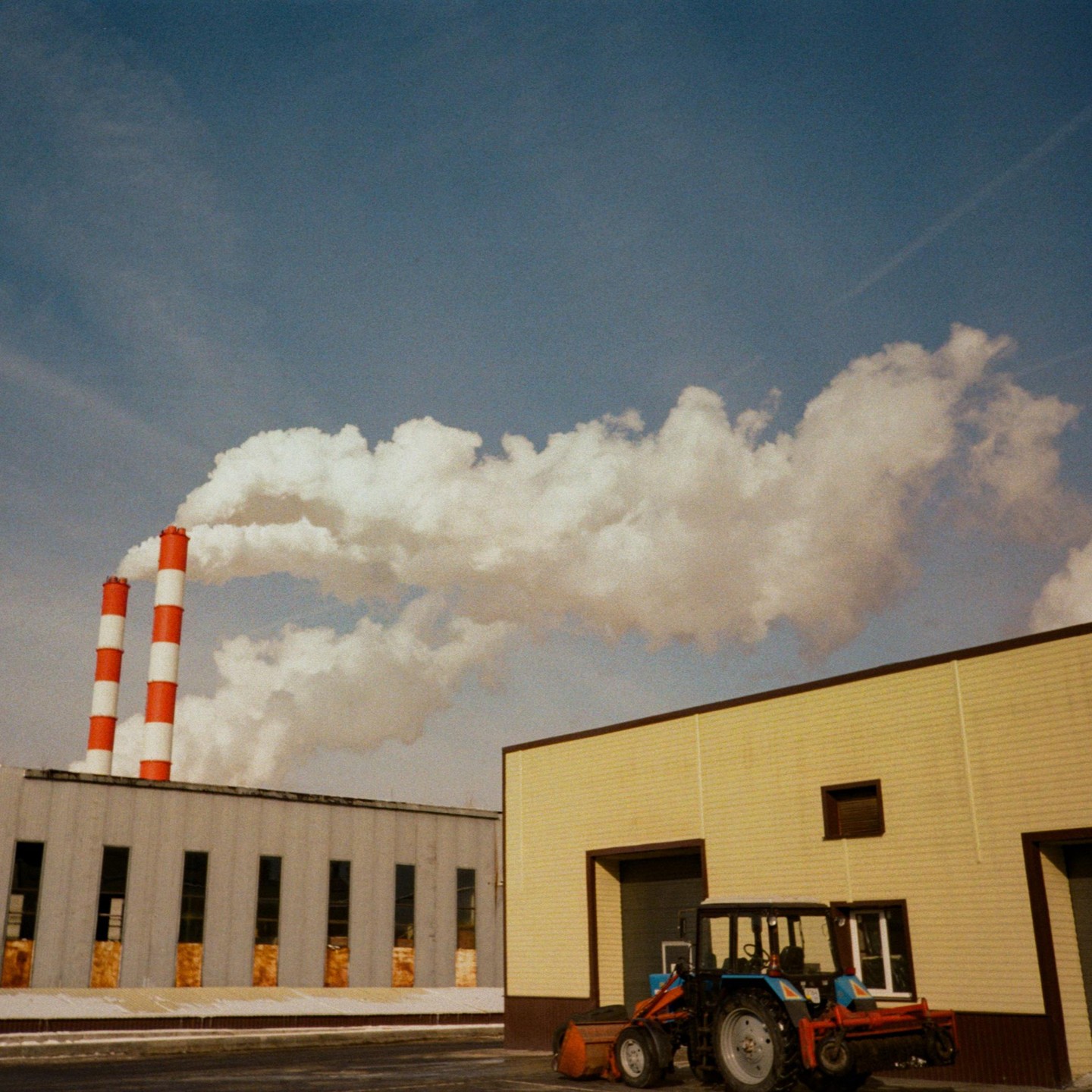 Two red and white striped smokestacks emit a large cloud of smoke next to a yellow building and a tractor.