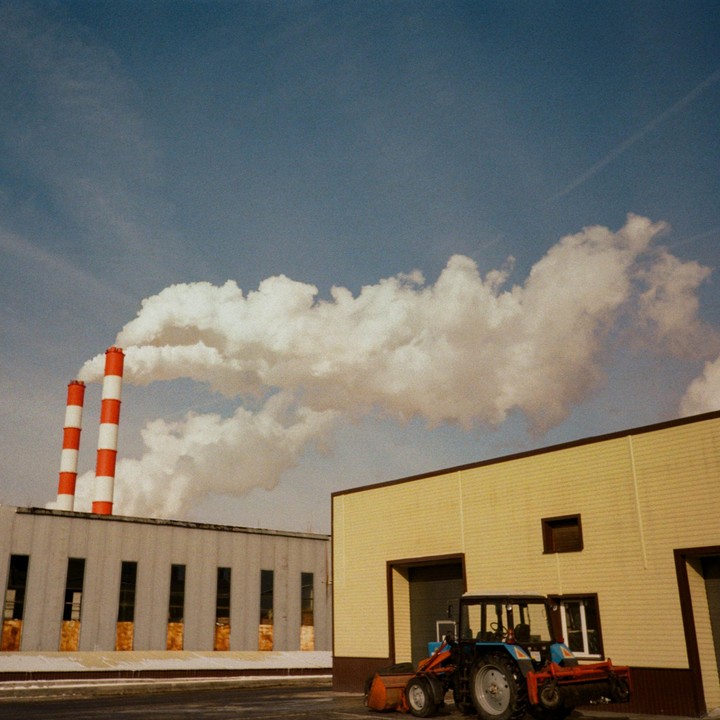 Two red and white striped smokestacks emit a large cloud of smoke next to a yellow building and a tractor.