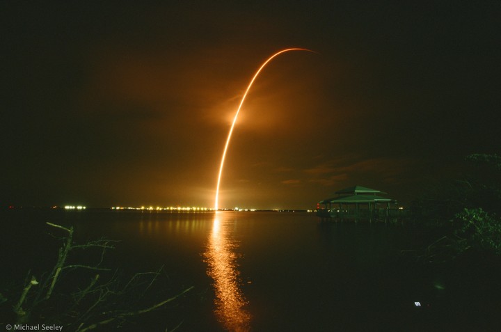 A long exposure captures the fiery arc of a rocket launch over a body of water at night, with a building on stilts visible on the right.