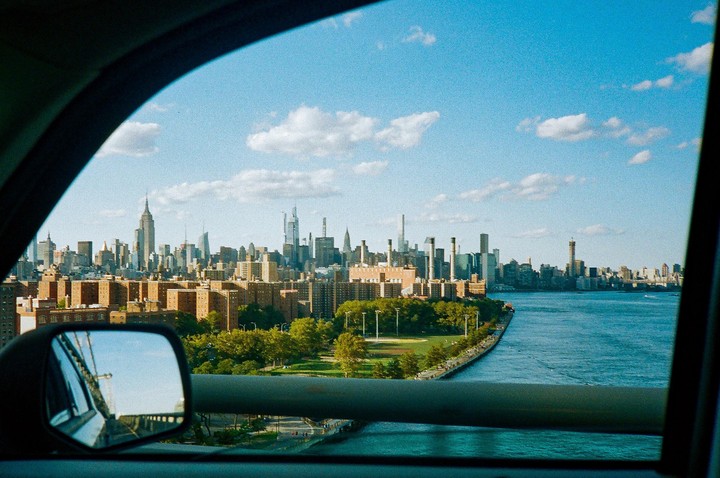 A view of the New York City skyline and a river from inside a vehicle, with a side mirror reflecting a bridge.
