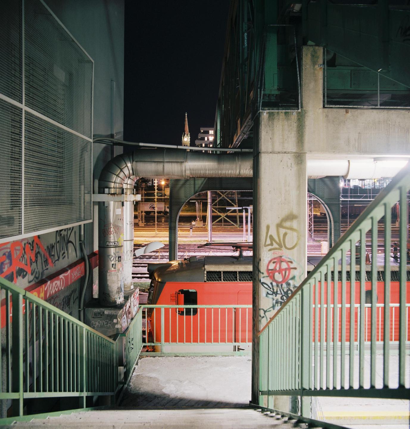 A red train is visible through a concrete structure with green railings and graffiti at night.