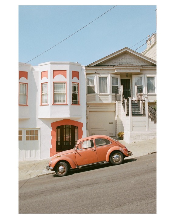 A peach-colored car is parked on a steep street in front of two houses.