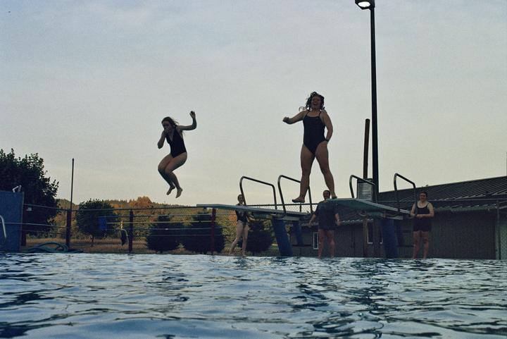 A woman jumps from a diving board into a swimming pool while other women stand on or near the diving board.