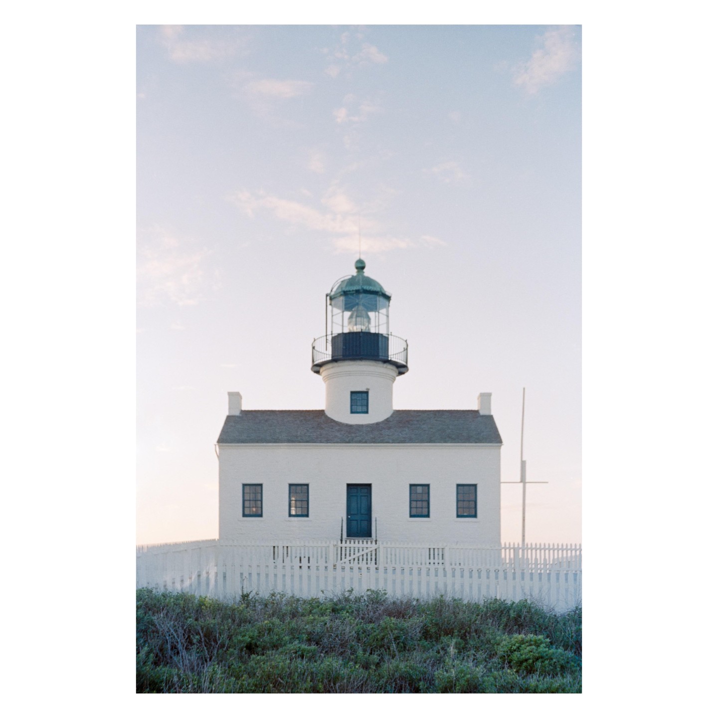 A white lighthouse with a dark blue door and windows stands behind a white picket fence and green bushes.