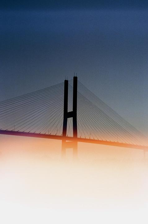 A cable-stayed bridge is silhouetted against a blue sky with an orange light leak across the bottom.