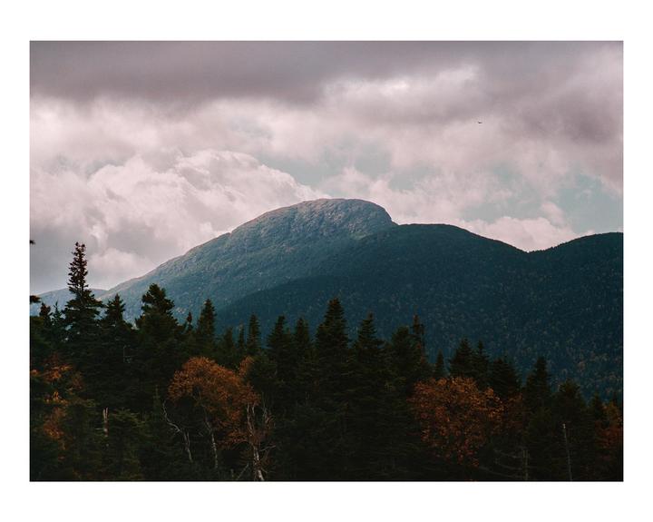 A mountain range covered in trees with some autumn foliage is visible under a cloudy sky.