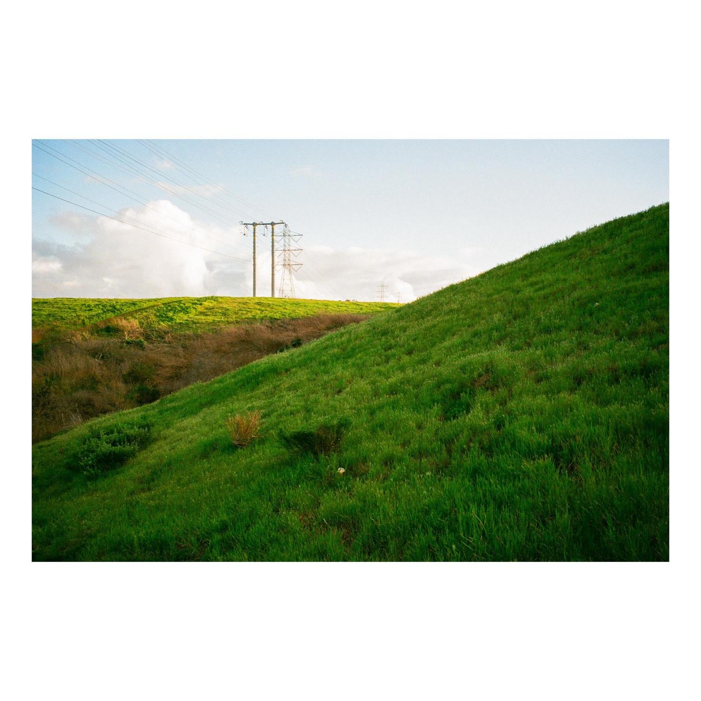A green hillside slopes upwards with power lines and transmission towers visible in the distance under a cloudy sky.
