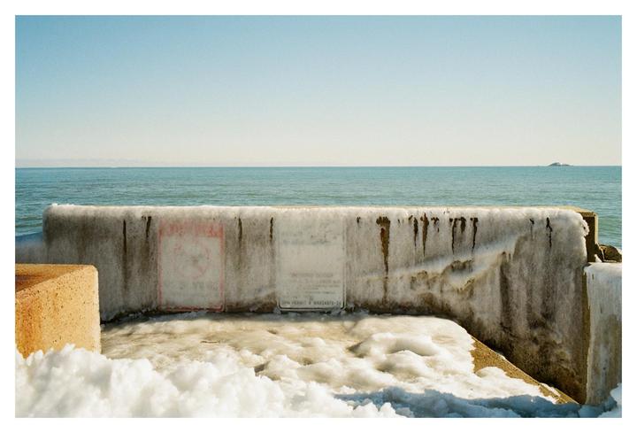 A concrete barrier covered in ice and snow stands before a body of water under a clear sky.