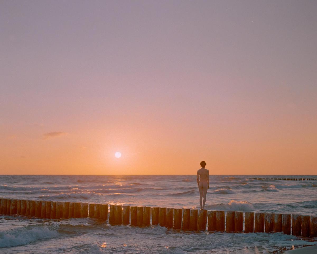 A woman stands on a wooden pier looking out at the ocean during sunset.