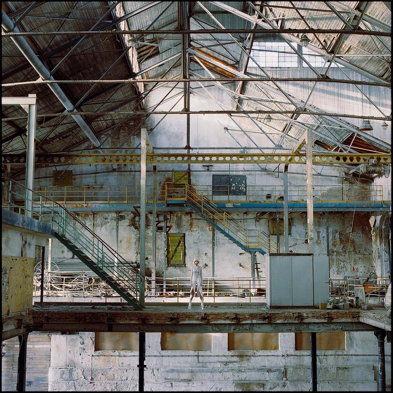 A woman stands on a platform in a large, dilapidated industrial building with exposed metal beams and staircases.