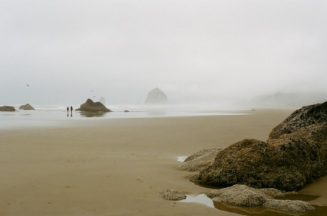 Two people walk on a sandy beach with large rocks and a hazy ocean in the background.