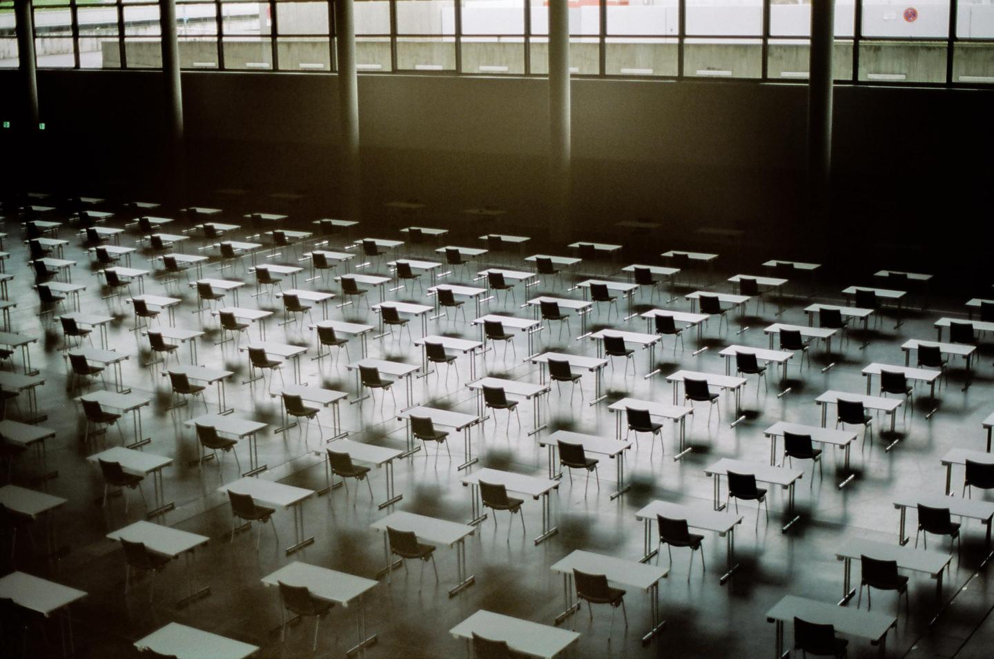 An empty examination hall is filled with rows of desks and chairs, illuminated by light from large windows.