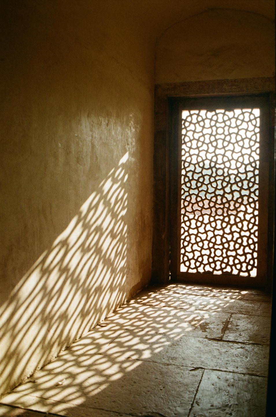 Sunlight streams through a patterned window, casting intricate shadows on the wall and floor of a room.