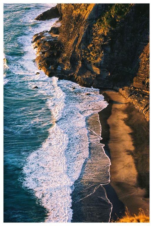 Waves crash against a rocky cliff and sandy beach during golden hour.