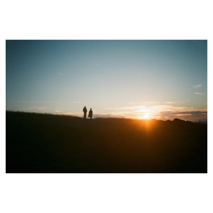 Two people stand silhouetted on a grassy hill at sunset.