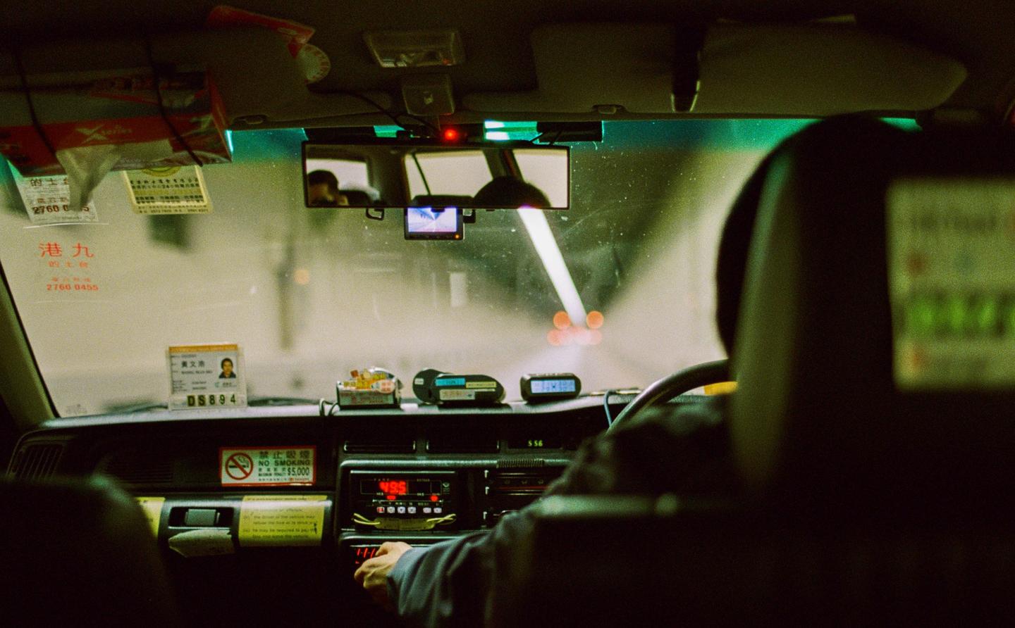 The interior of a taxi is visible from the back seat, with the driver's headrest and dashboard in the foreground and a tunnel visible through the windshield.