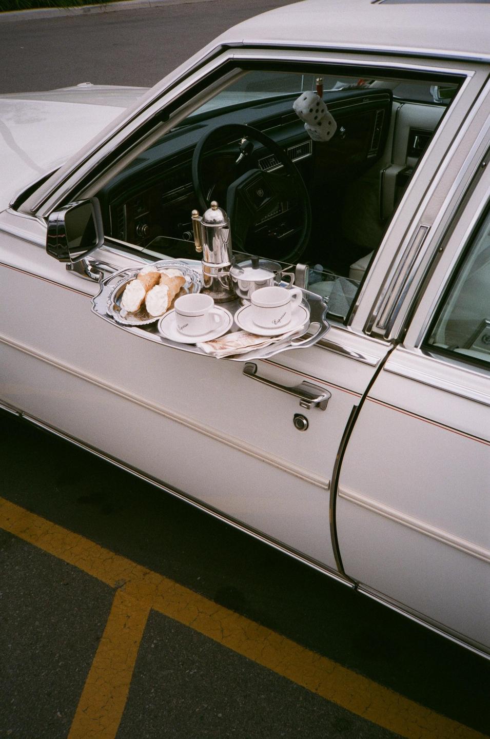 A silver tray with coffee cups and pastries is placed on the open window of a white car.