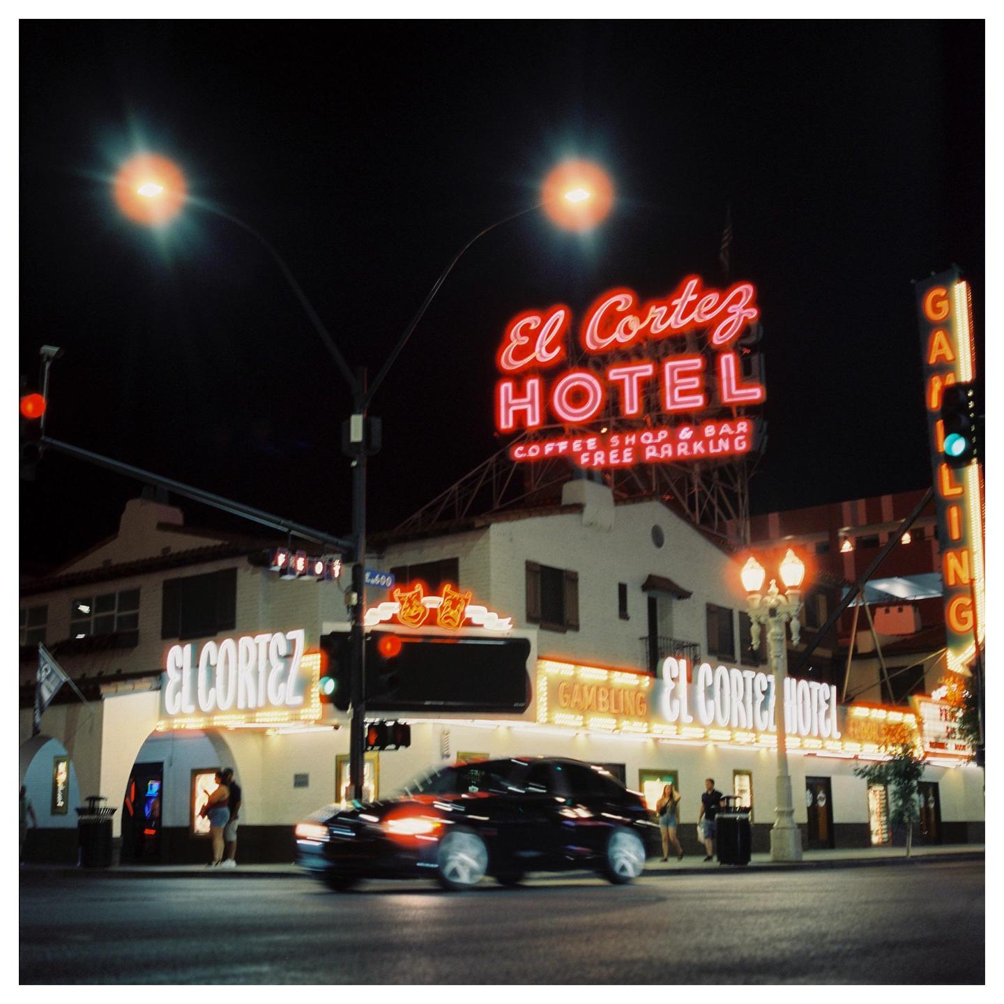 A black car with motion blur drives past the El Cortez Hotel at night, illuminated by neon signs and streetlights.