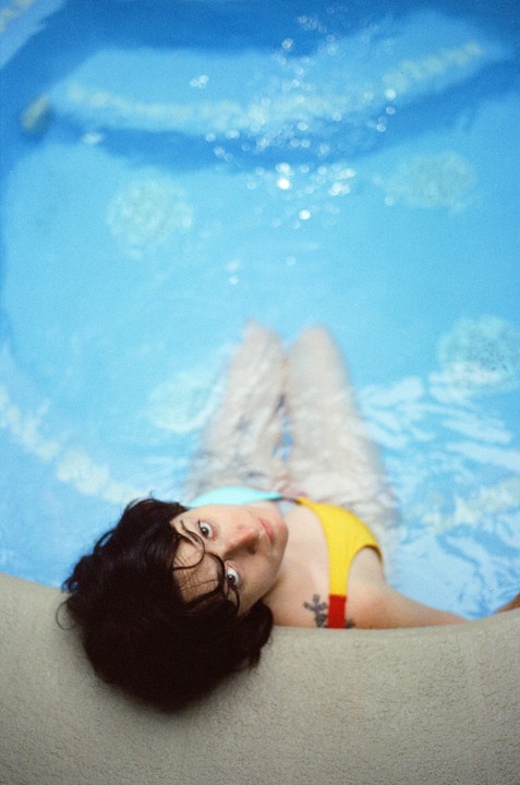 A woman with dark hair and glasses floats in a blue hot tub, looking up at the camera.
