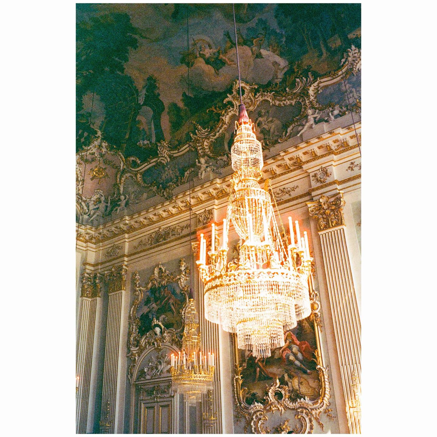 A large, ornate crystal chandelier hangs from a highly decorated ceiling in a grand room.
