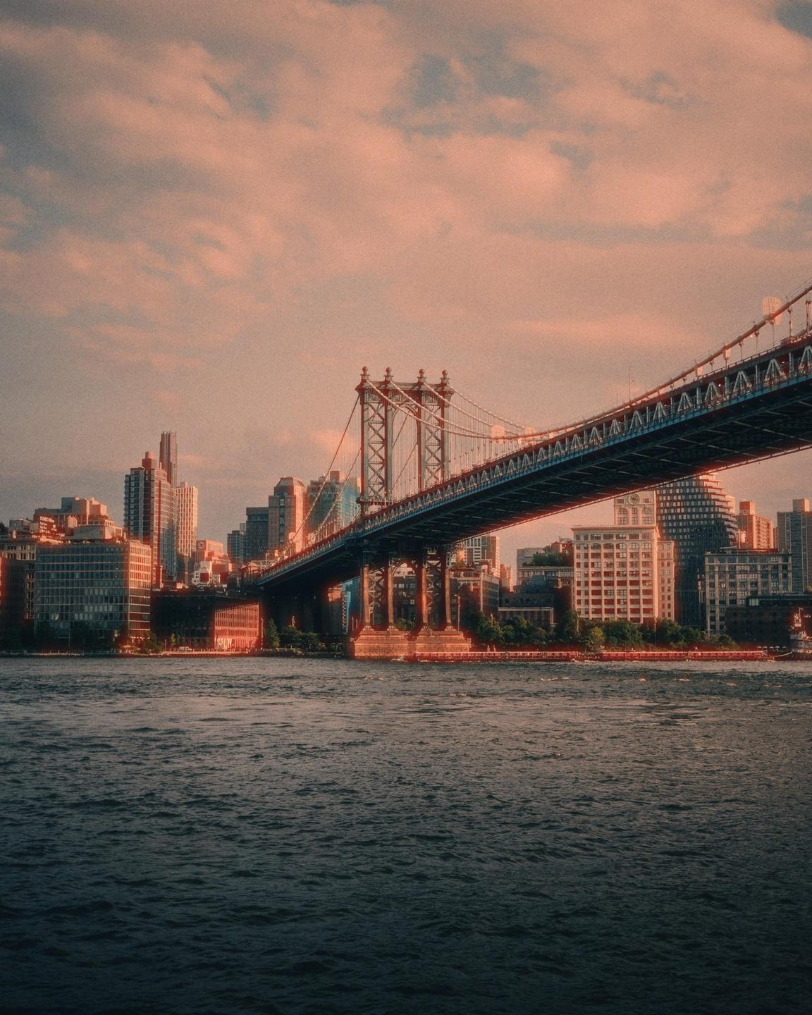 The Manhattan Bridge spans a body of water with a city skyline in the background under a cloudy sky.