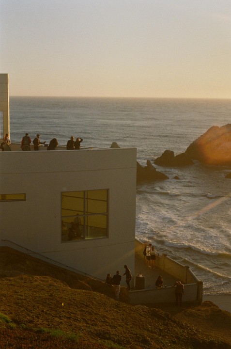 A group of people stand on the roof and balcony of a building overlooking the ocean at sunset.