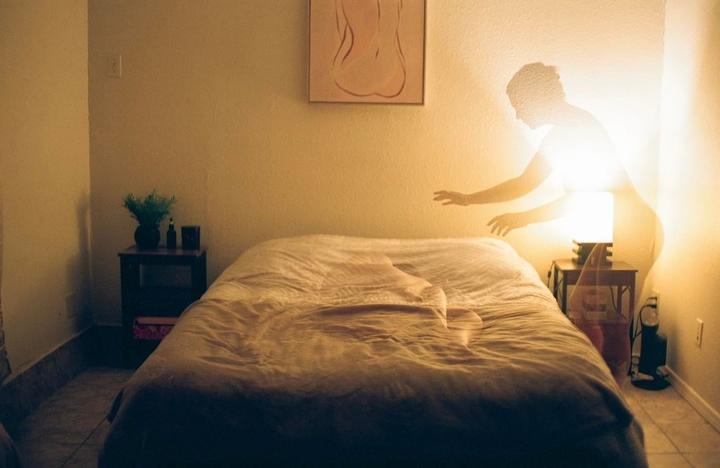 A glowing figure stands next to a bed with a faint impression of a body on the sheets.