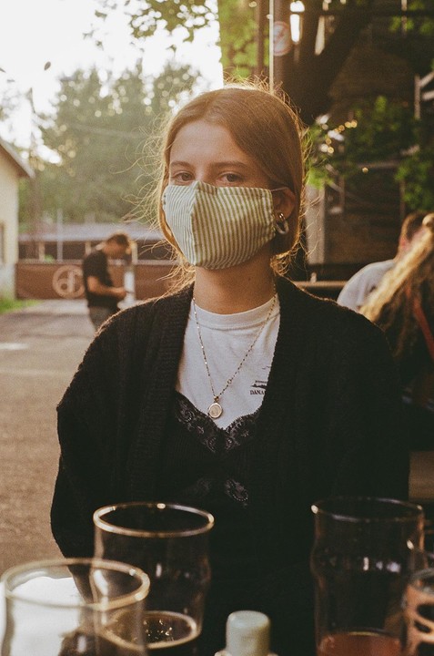 A woman wearing a striped face mask and a necklace sits at a table with drinks.