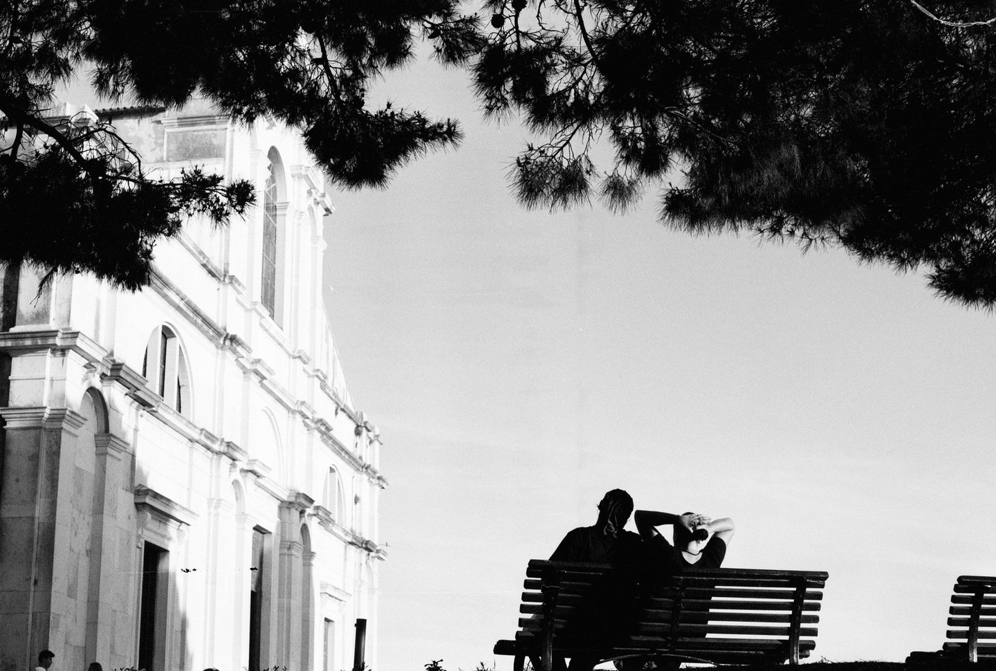 Two people sit on a bench looking at a white building with a tree branch in the foreground.