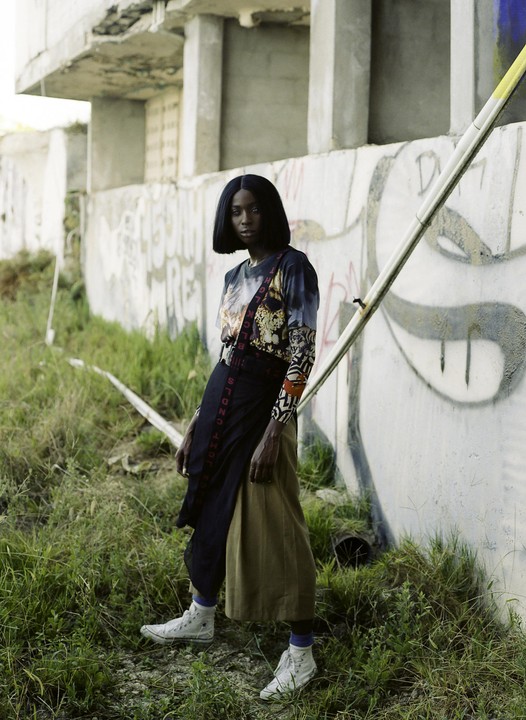 A woman stands in tall grass next to a concrete wall with graffiti.