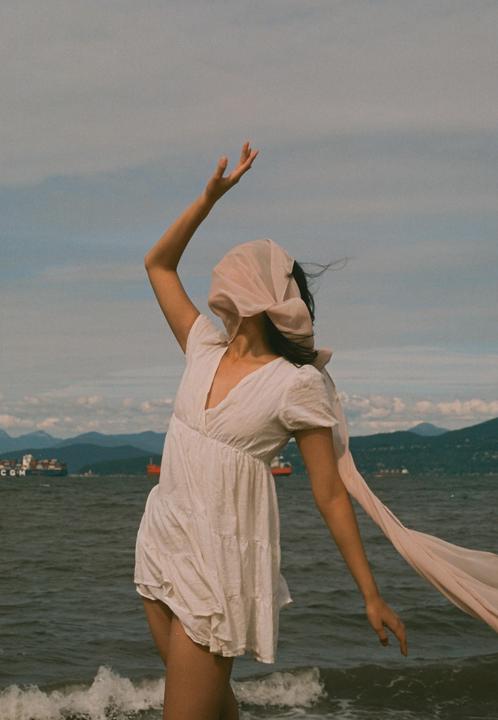 A woman in a white dress with a scarf covering her face stands by the ocean with mountains and ships in the background.